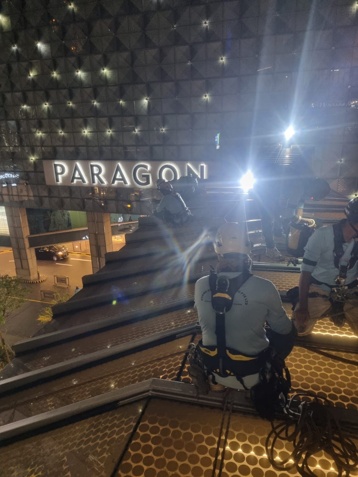 Rope-access technicians abseiling above an illuminated Orchard Road mall entrance at night